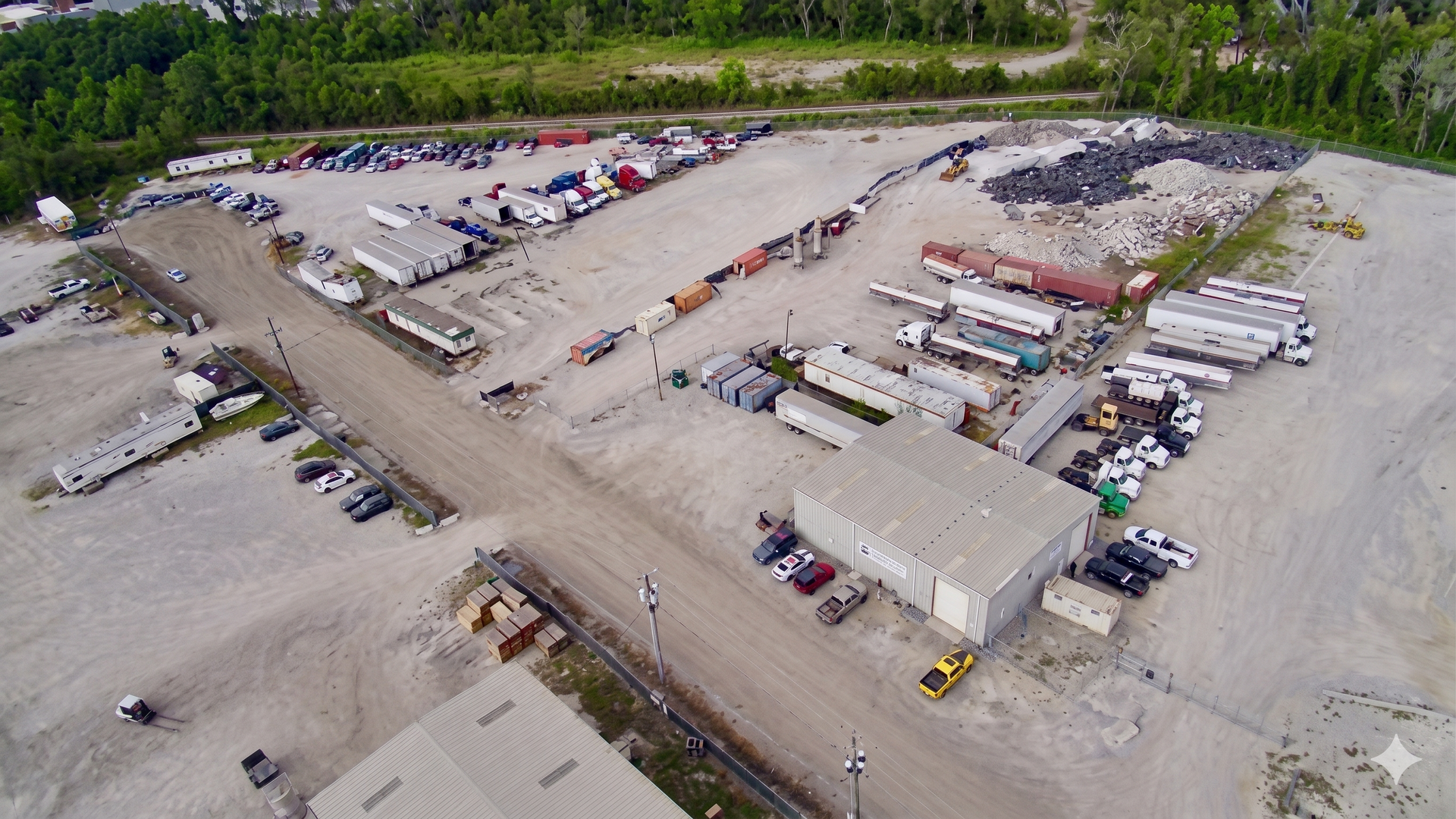 Aerial view of a gravel industrial yard with parked trucks, trailers, storage containers, and trees nearby
