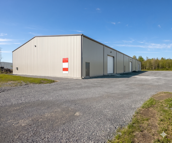 Large beige metal warehouse beside a gravel lot under a clear blue sky