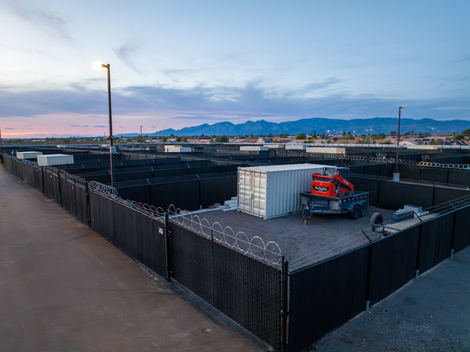 Construction site with black fencing, red excavator, and shipping container at dusk with mountains in the background