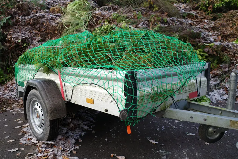 Small utility trailer covered with green netting, parked on a leaf-covered ground outdoors.
