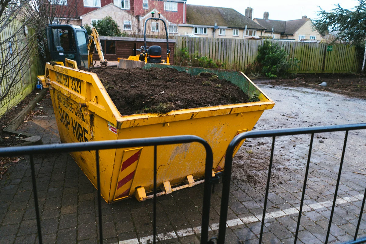 Yellow construction dumpster filled with soil and debris on a wet street, behind metal barriers