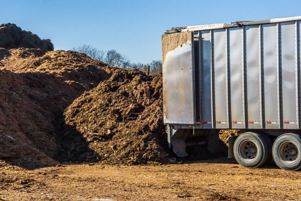 Large metal truck trailer parked beside a mound of wood chips in an outdoor yard