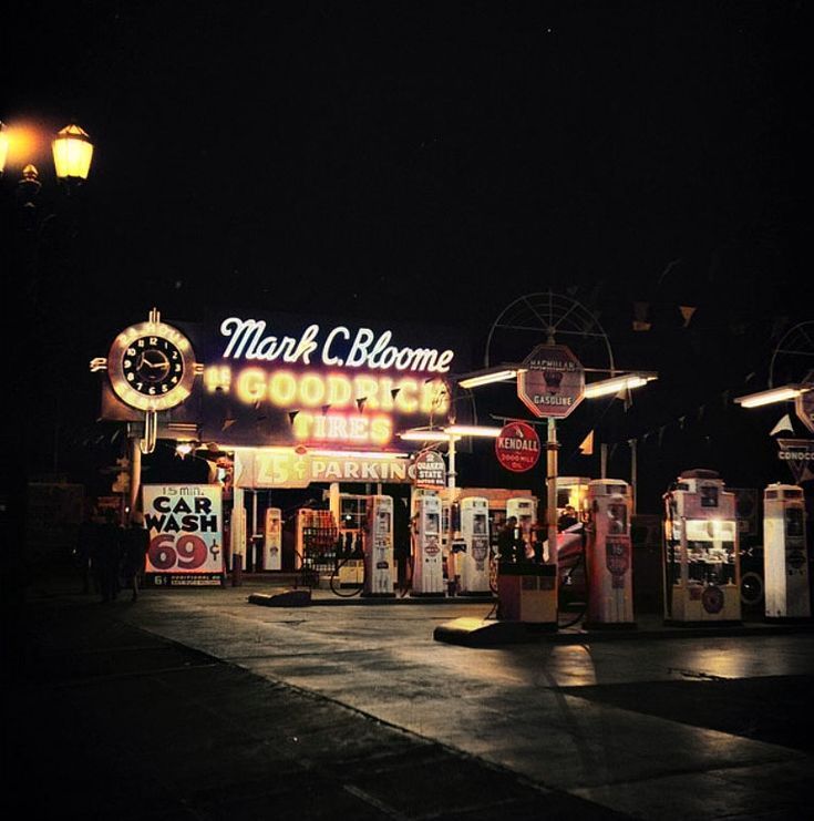 Nighttime vintage gas station lit with neon signs and antique pumps.