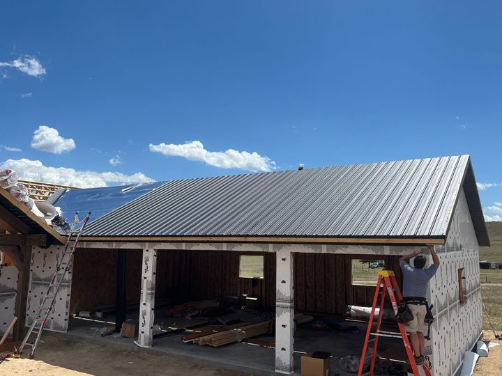 Partially built house with gray metal roof under a blue sky, workers and ladder in the foreground