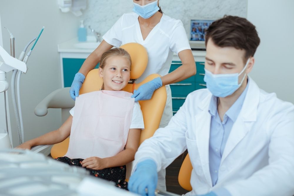 Dentist, assistant, and young patient in dental office. Dentist and assistant are masked and gloved. Patient is wearing bib.