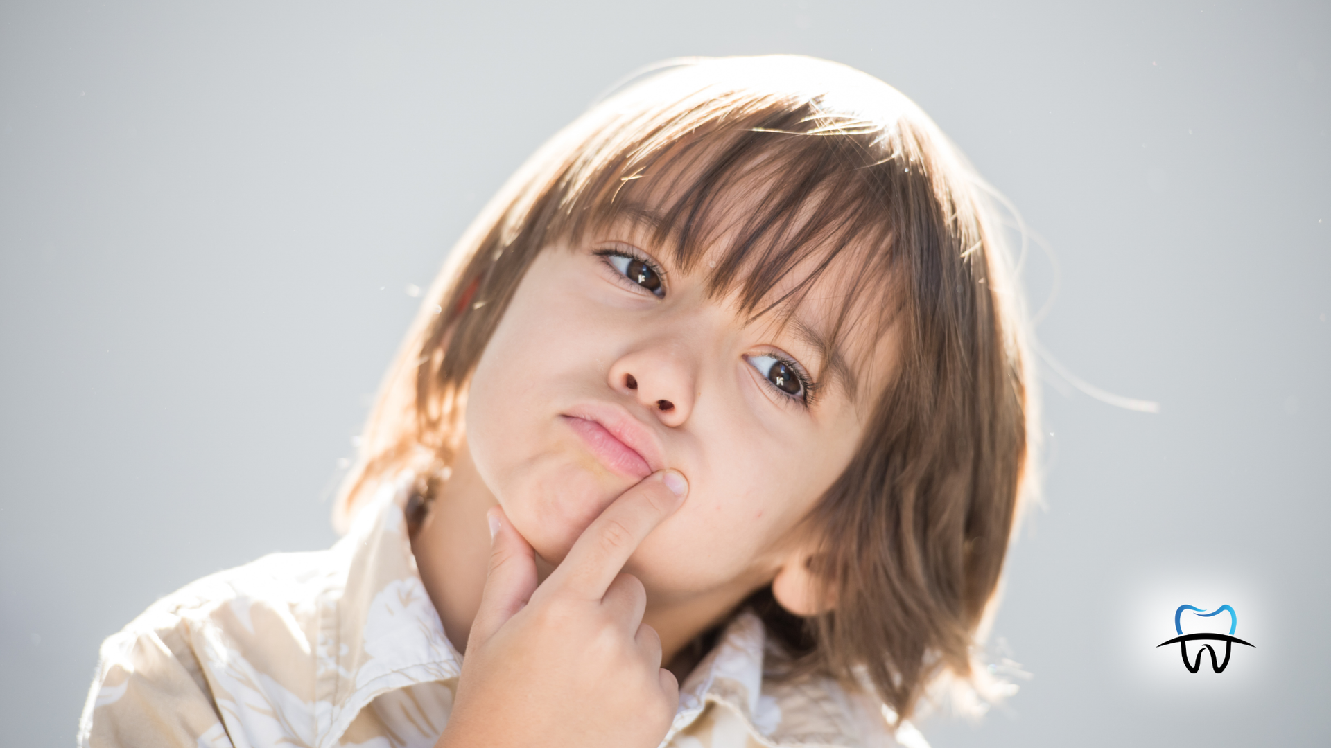 Boy with hand on chin, looking thoughtful, light shirt, gray background.