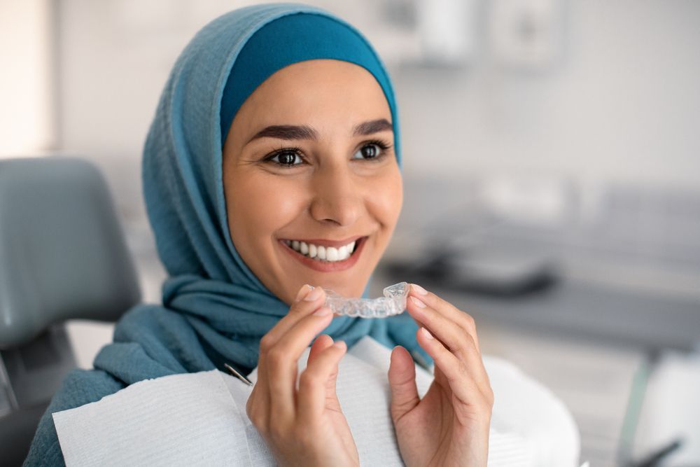 Woman in hijab holding clear aligner, smiling in dentist's office.