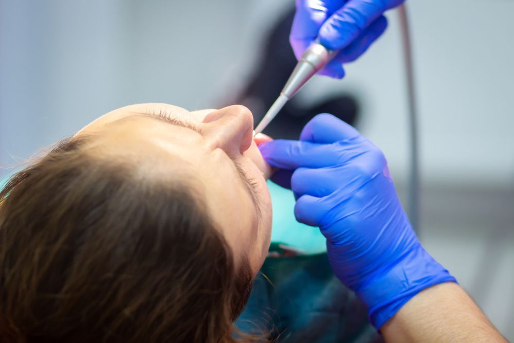 Dentist working on a patient's teeth with a drill. Blue gloves, dental office setting.