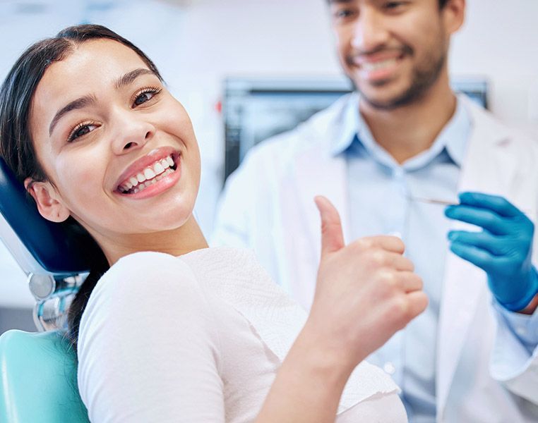 Woman in dentist chair smiles and gives thumbs up; dentist smiles in background.