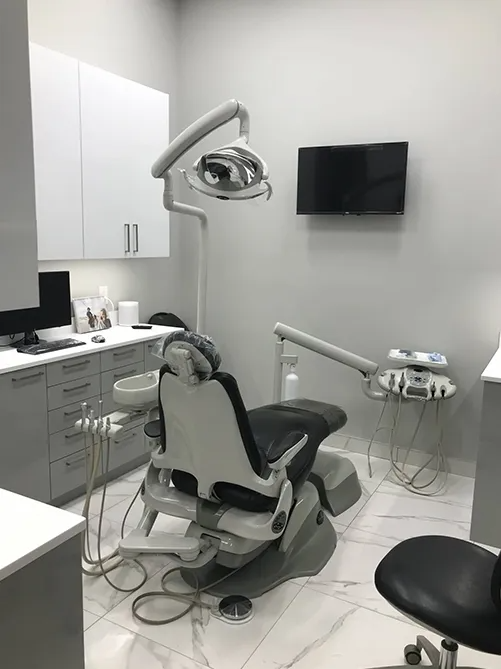 Empty dental exam room with chair, overhead light, cabinets, and wall-mounted TV.