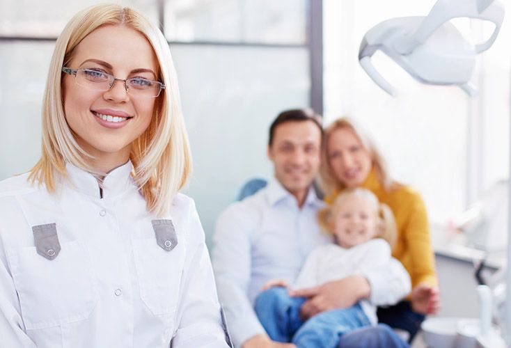 Dentist smiling at the camera, a family sitting in dental chairs in the background.