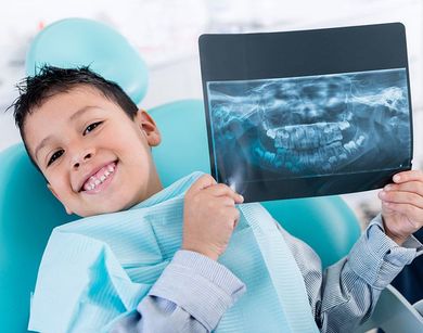 Boy in dental chair smiles, holding up a dental X-ray, turquoise and white setting.