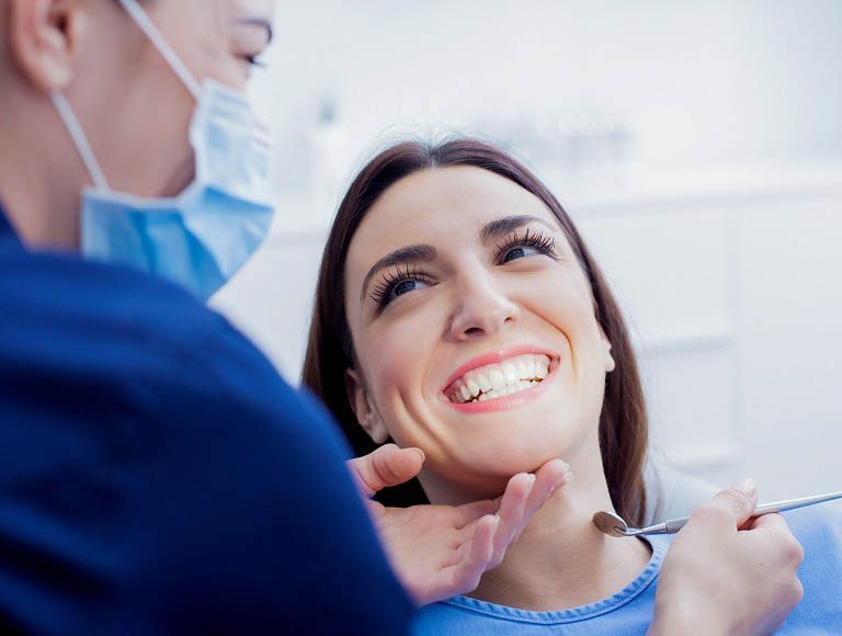 Woman smiling at dentist during a check-up; dentist in mask holding tools.