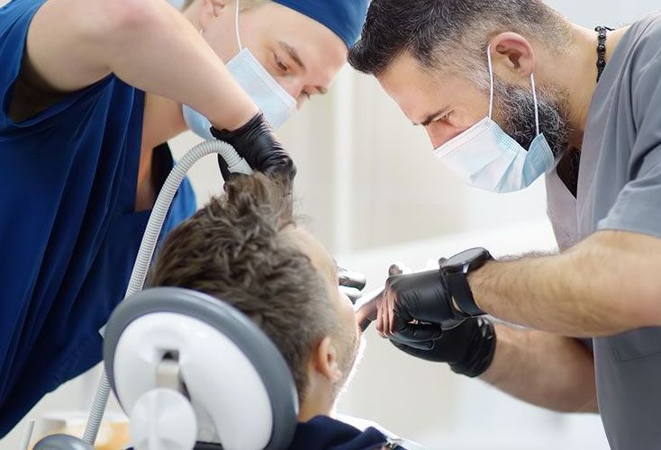 Two dental professionals, masked and gloved, tending to a patient's mouth; dental setting.