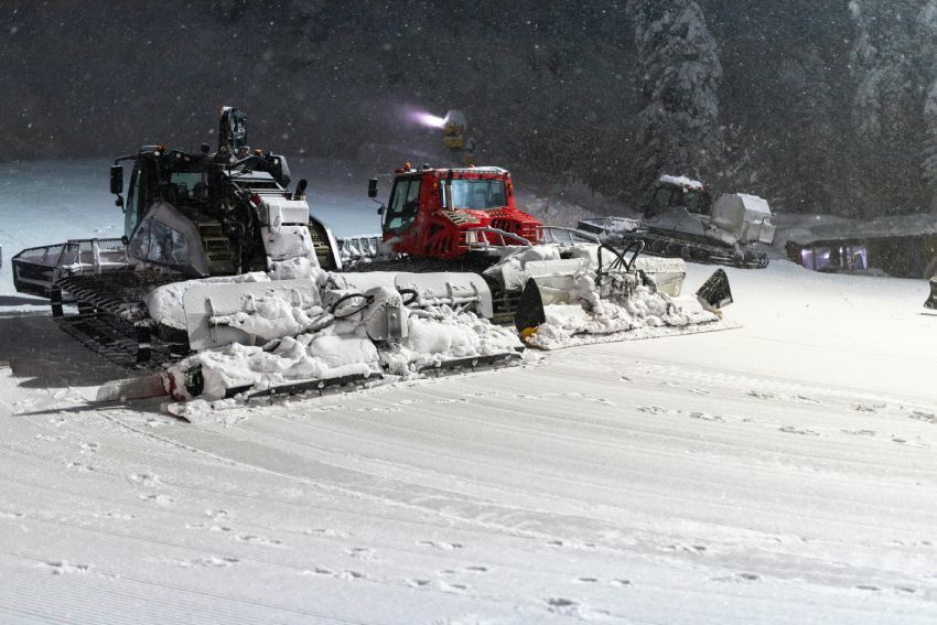 Snow groomers preparing ski slope at night during snowfall with fresh corduroy trails and forest backdrop for resorts nightly