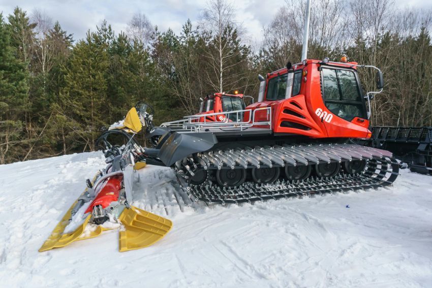 A large snow groomer with wide tracks and snow plowing blades, parked on a snowy mountain slope, surrounded by trees.