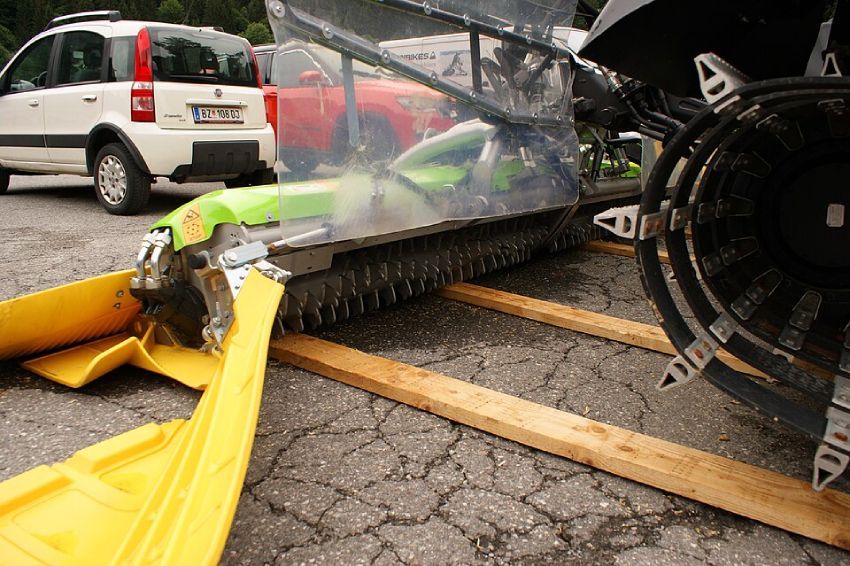 A snow grooming machine’s plow attachment, with its yellow blades and tracked wheels, resting on wooden planks.