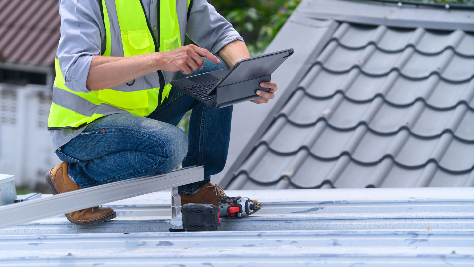 Worker in a neon safety vest using a tablet on a rooftop beside gray roof tiles.