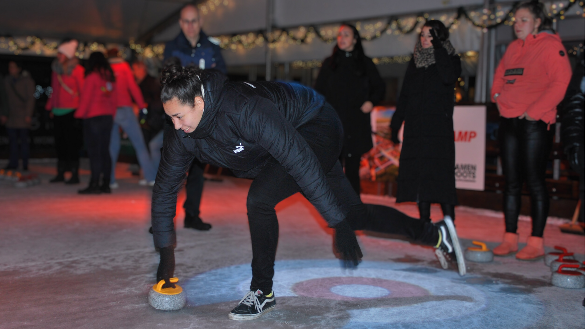 Iemand schuift een curlingsteen over het ijs, anderen kijken toe in een winterse omgeving.