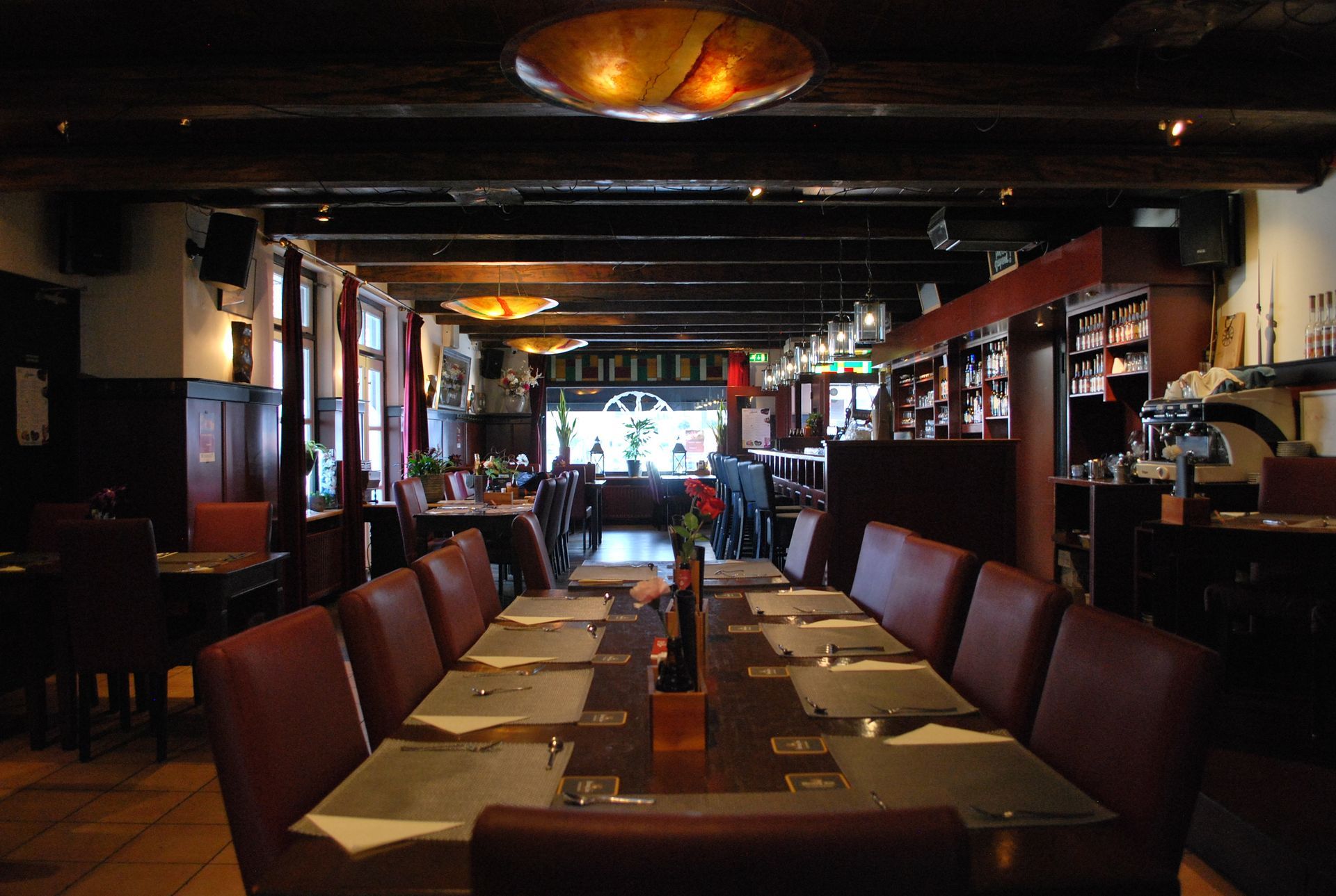 Restaurantinterieur met lange eettafels, rode stoelen en zichtbare houten balken.