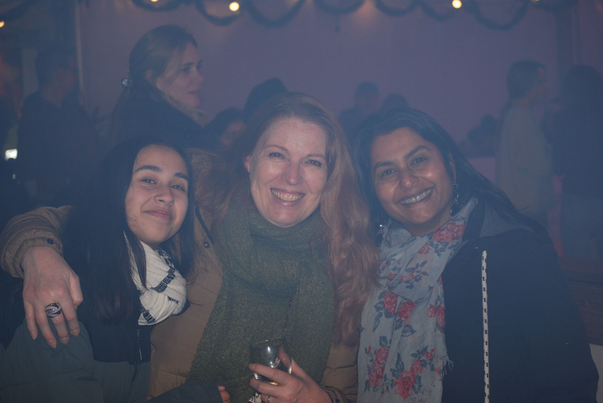 Drie vrouwen glimlachen, arm in arm, in een schemerig verlichte binnenruimte.