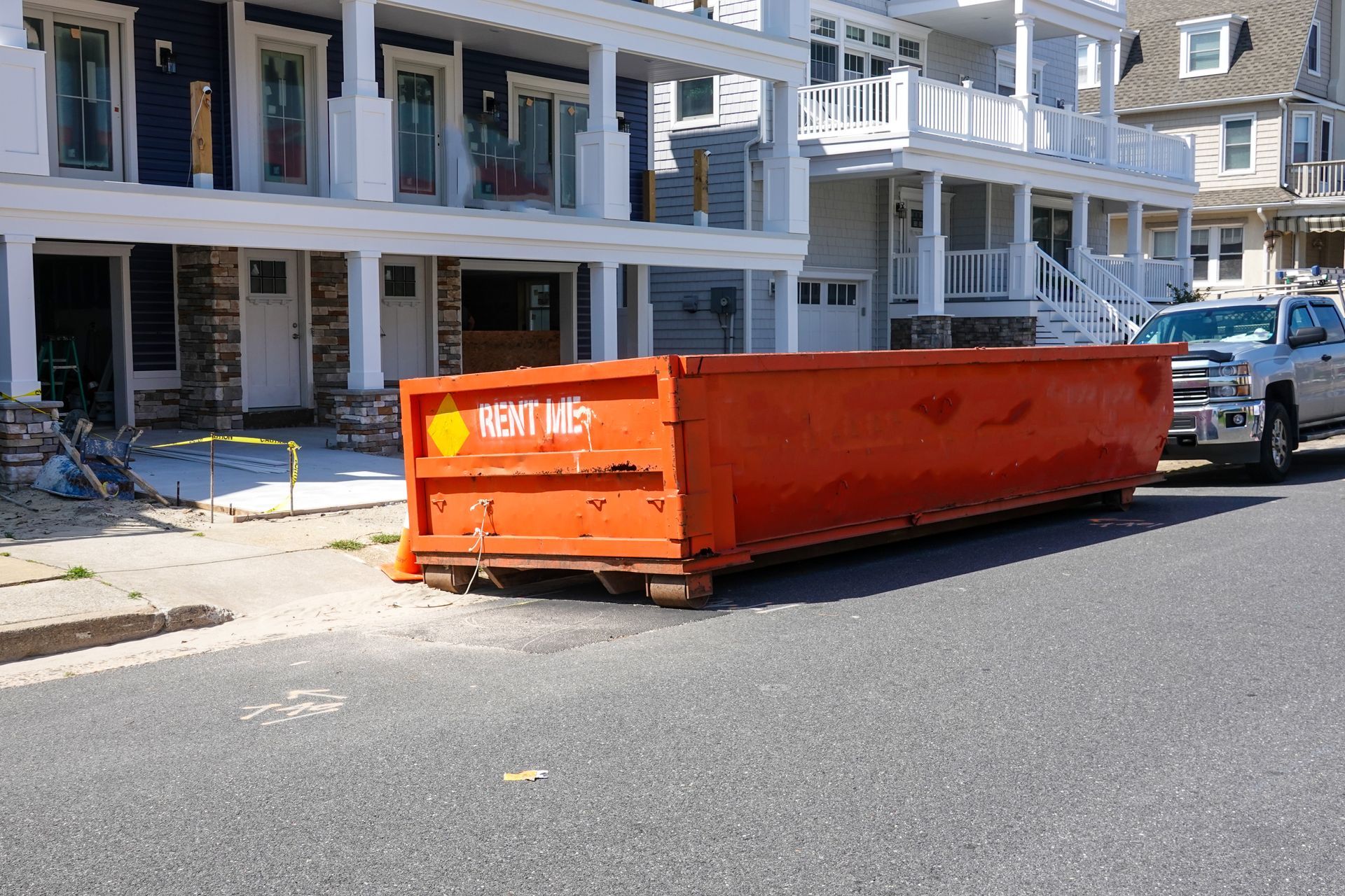 An orange dumpster on a street in front of a truck on a sunny day.