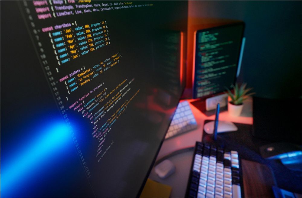 Dual monitors displaying code in a dark, neon-lit workspace with a mechanical keyboard and a small plant.