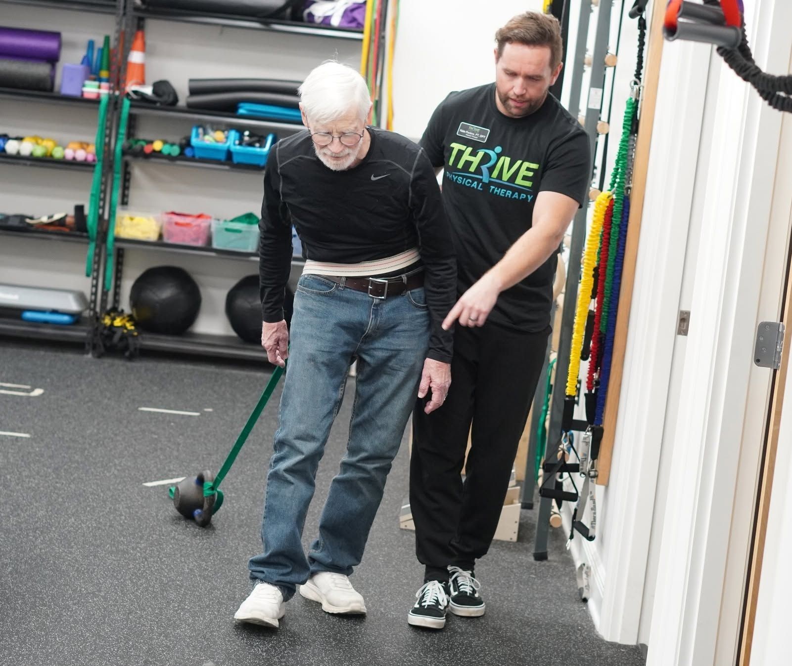 Older person walking with assistance in a gym, guided by a trainer beside them.