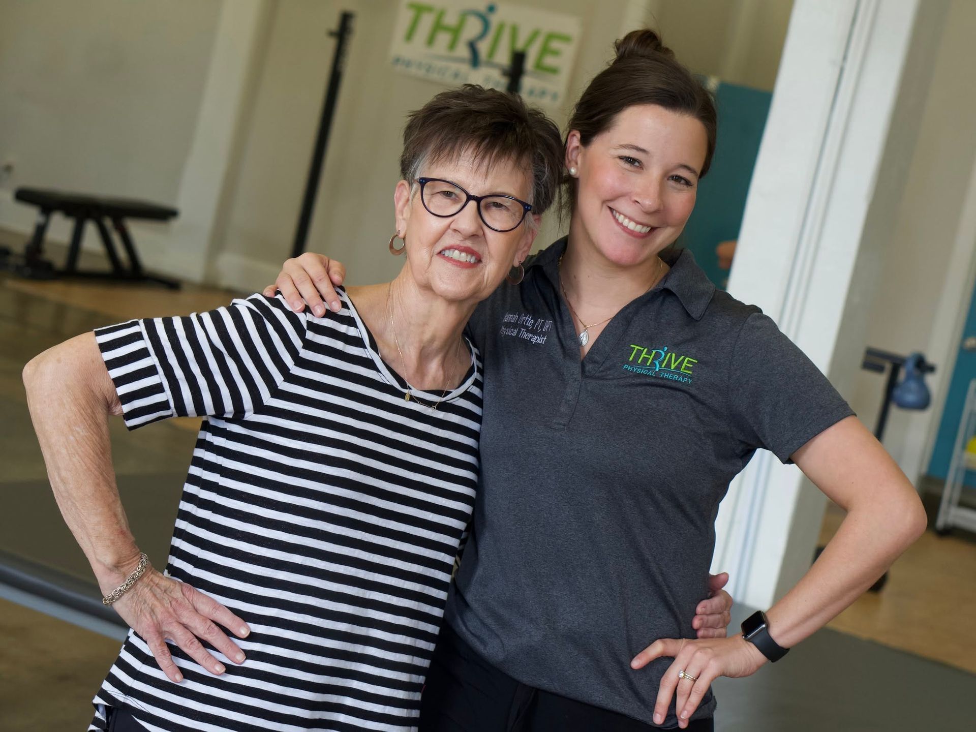 Two women smiling with arms around each other in a bright office lobby, wearing striped and gray polo shirts.