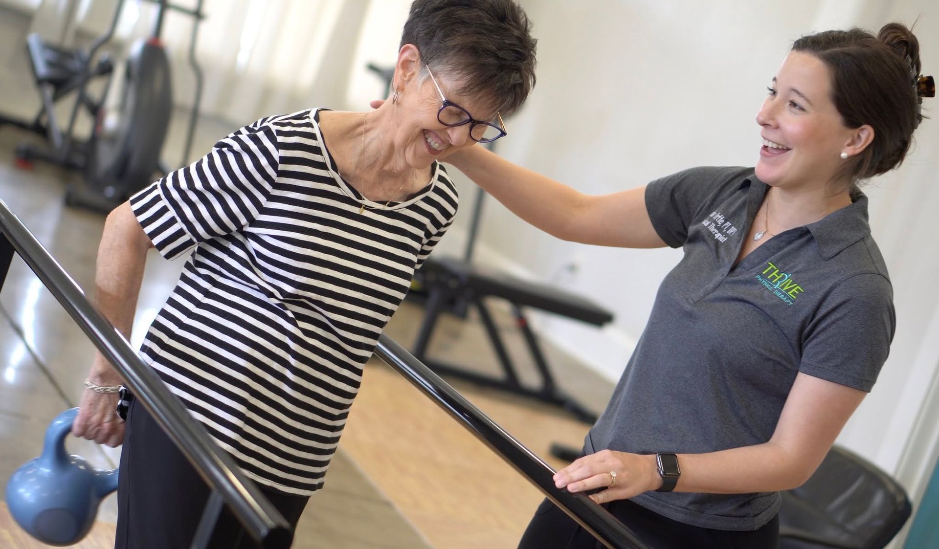Trainer assisting a woman on gym equipment, both smiling in a bright fitness center