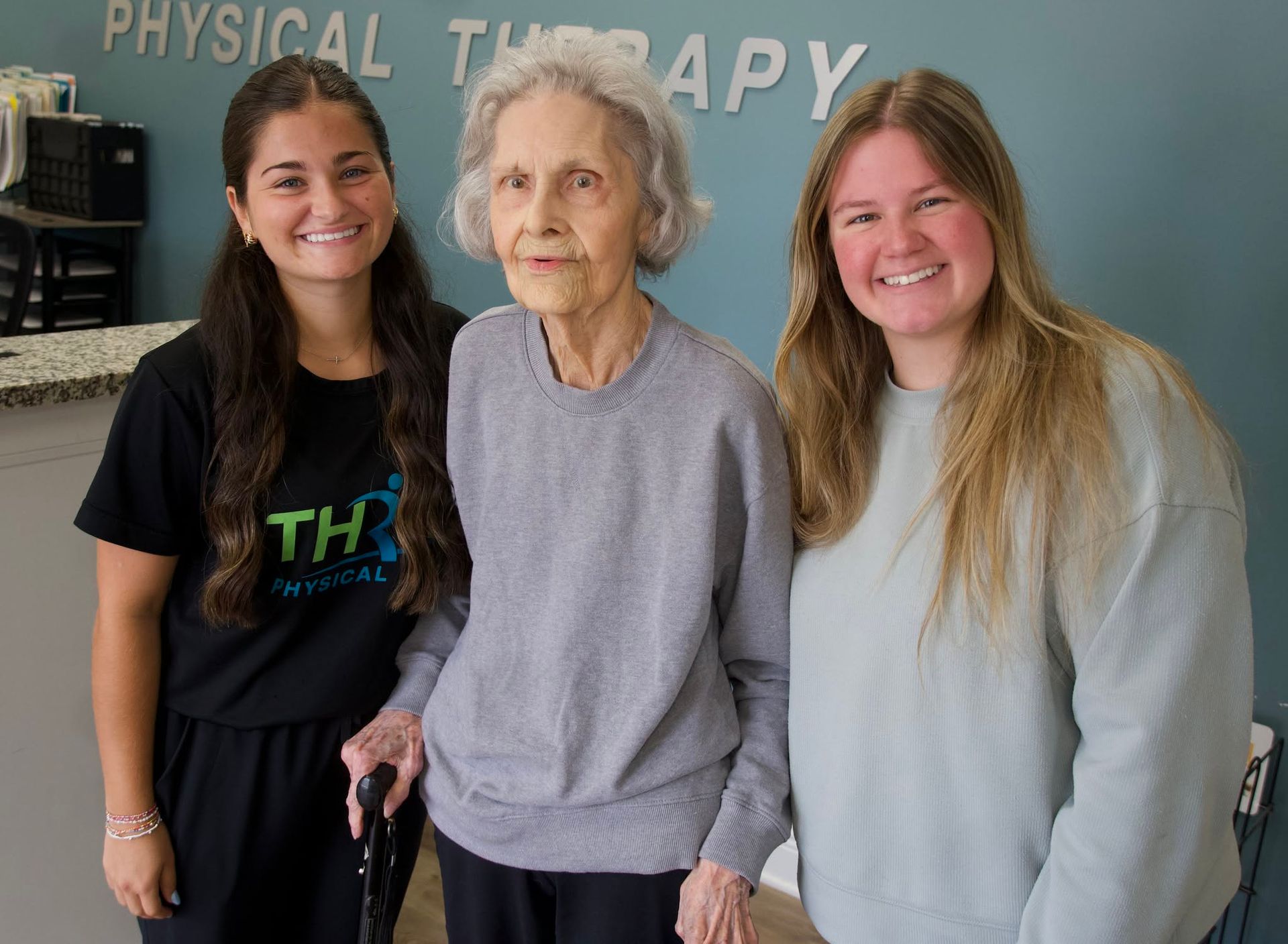 Three women standing together in a physical therapy clinic, smiling at the camera.