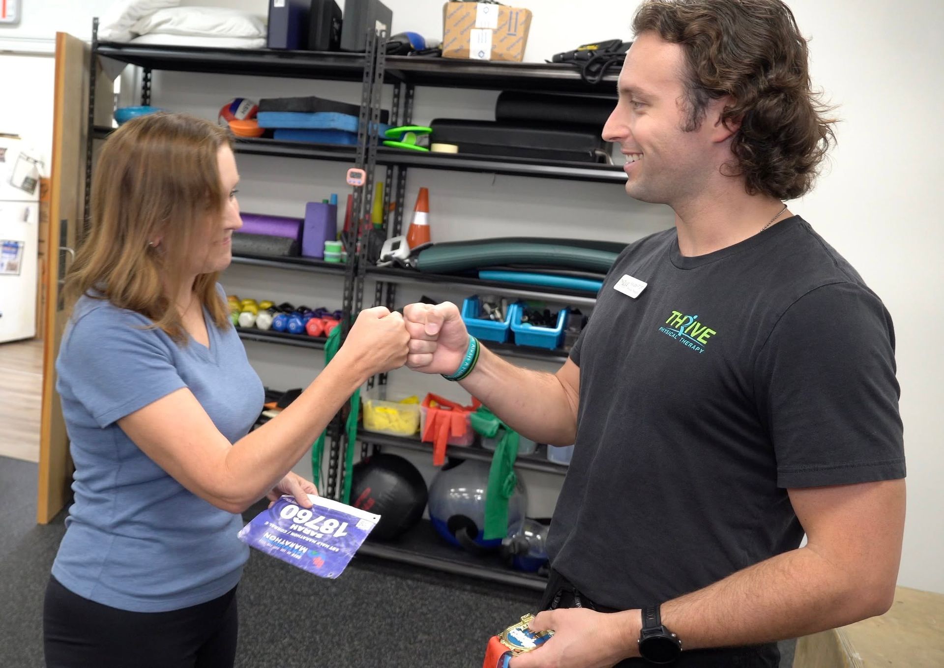 Two people smiling and shaking hands in a gym, with exercise equipment on shelves behind them