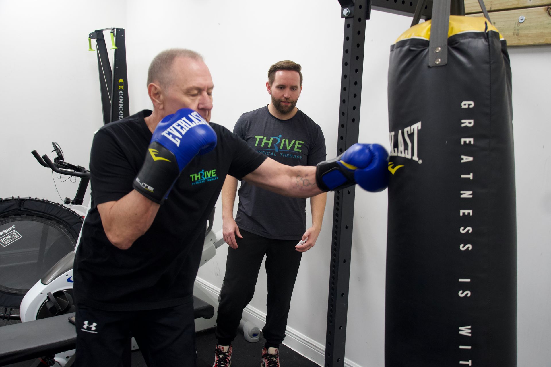 Man punching a heavy bag while another watches in a gym, both wearing boxing gloves and black shirts