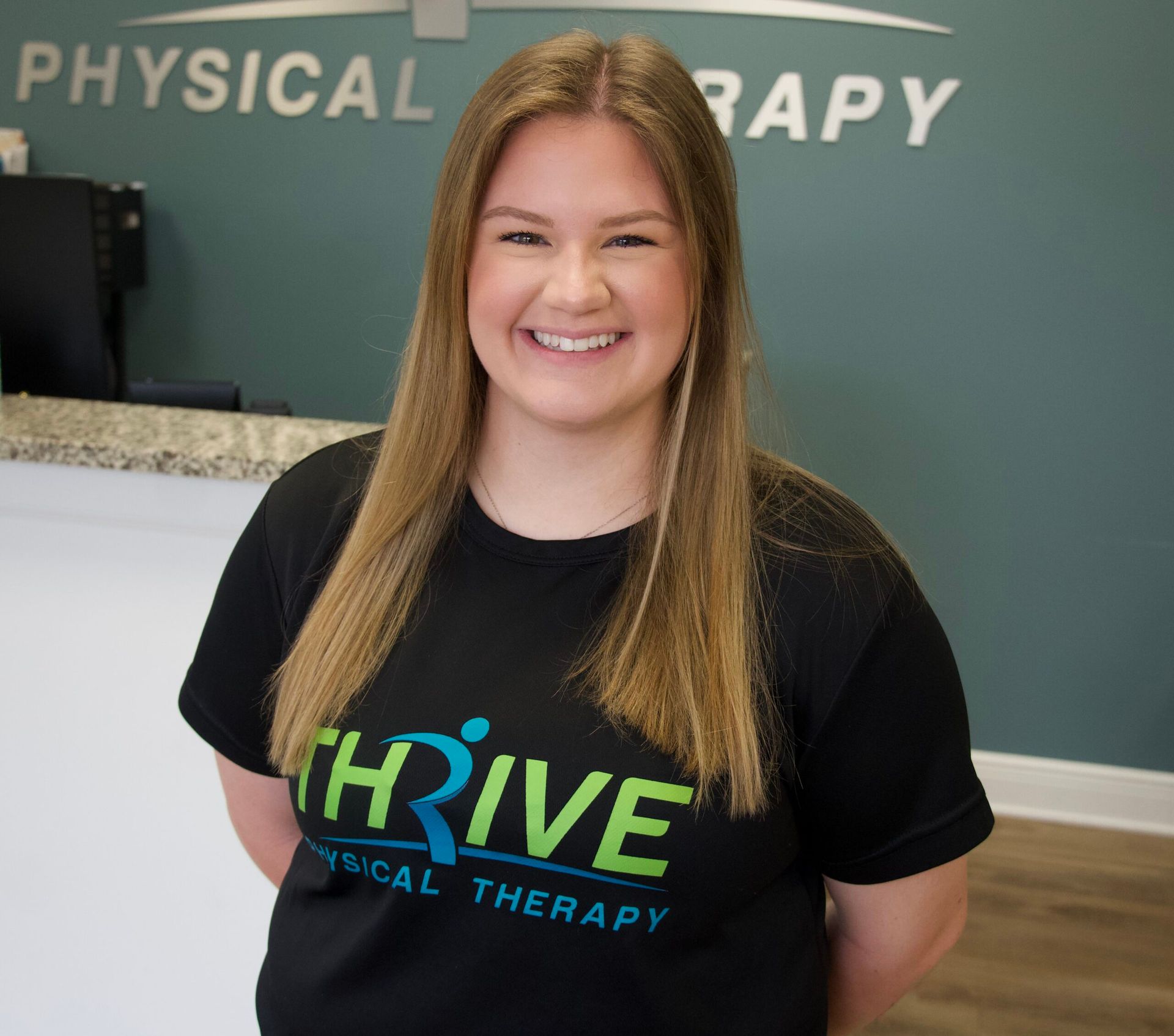 Woman in black THRIVE Physical Therapy shirt smiling in a clinic lobby.