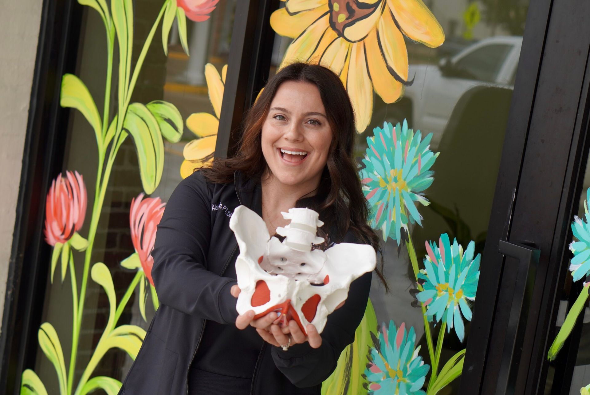 Woman holding a white baby goat in front of a colorful flower mural and smiling at the camera