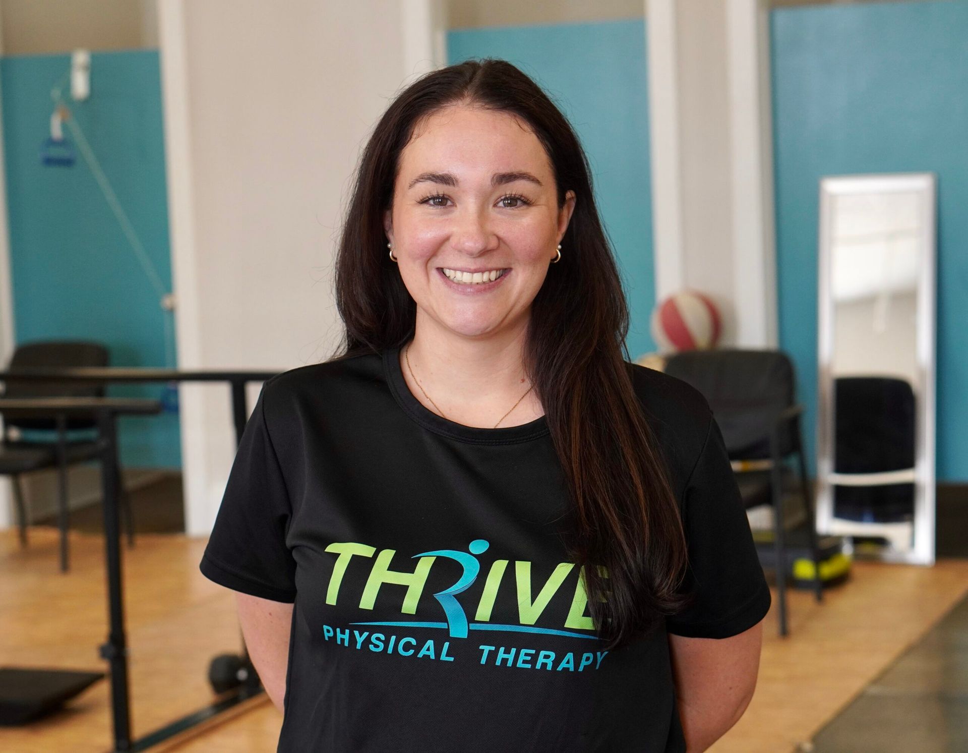 Woman smiling in a gym, wearing a black THRIVE Physical Therapy shirt.