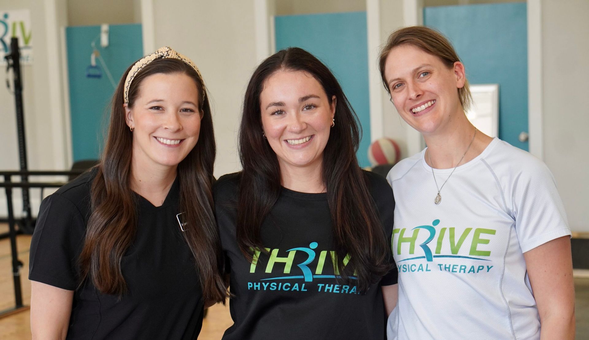 Three women standing together indoors, smiling in Thrive Health and Wellness shirts