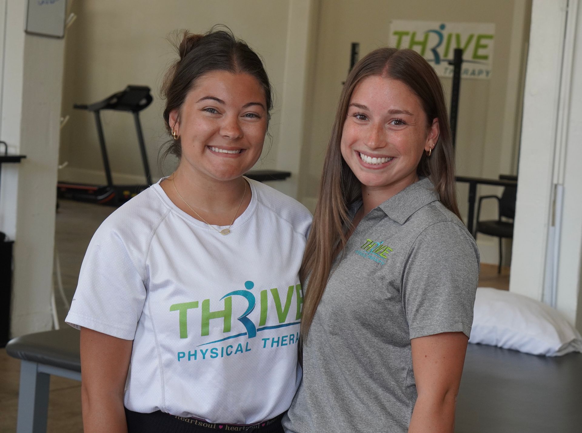 Two smiling physical therapists standing in a clinic, one in a white shirt and one in a gray polo.