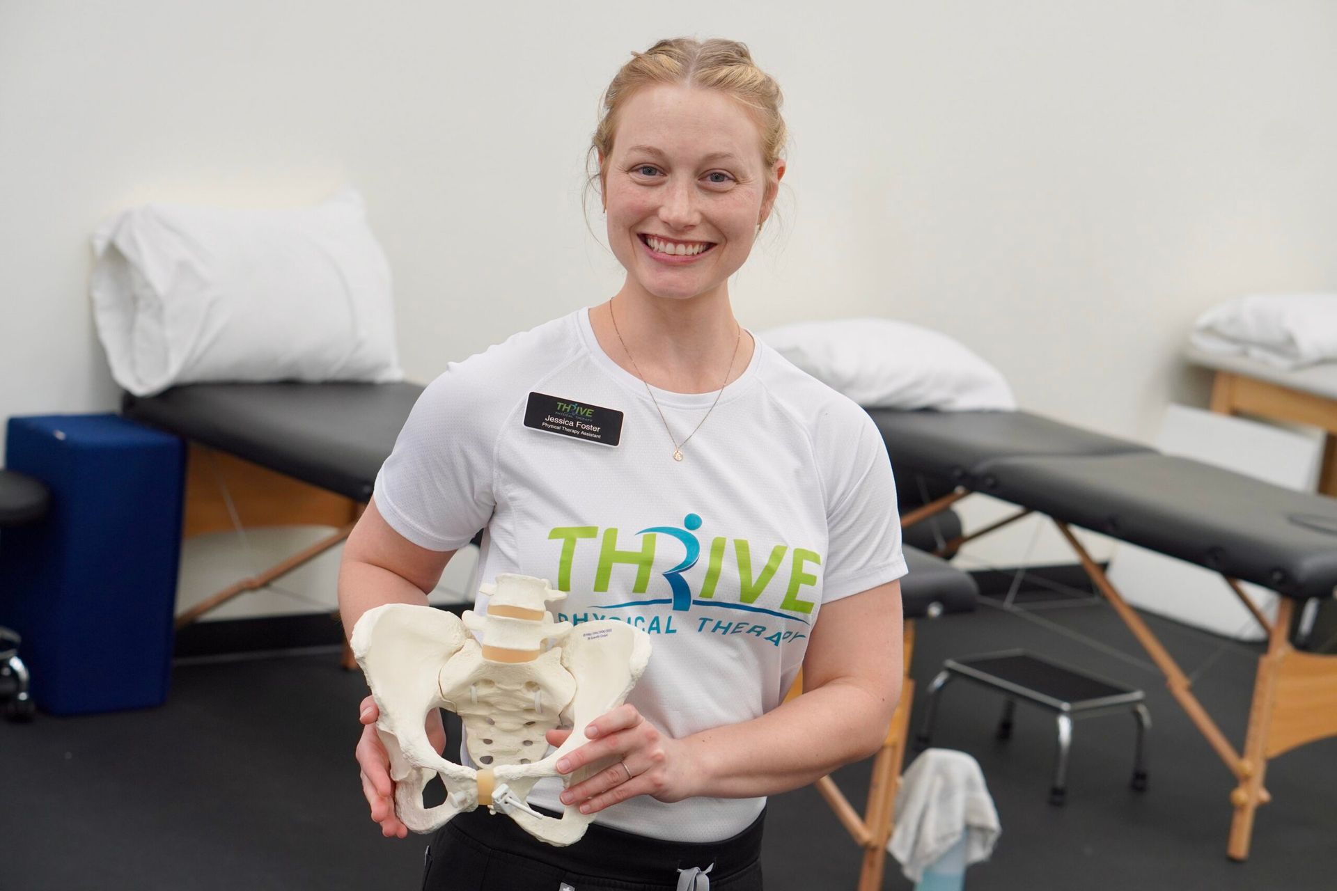 Smiling woman holding a pelvis model in a physical therapy room with treatment tables
