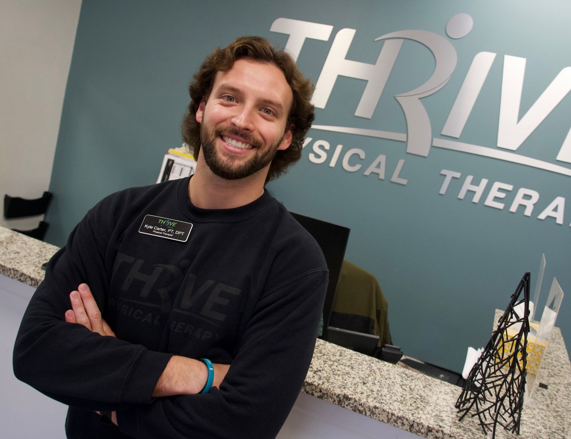 Smiling person with arms crossed in a physical therapy clinic lobby with THRIVE sign behind them