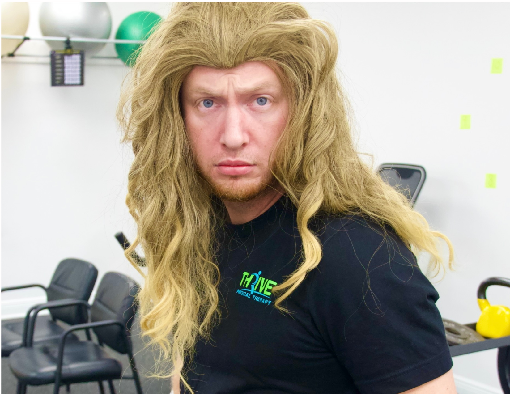 Man with long blond hair and black shirt in a gym, facing camera with a stern expression