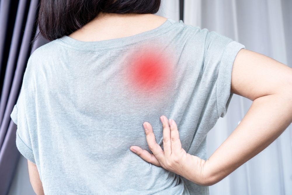 Person holding their back with a red pain spot on a gray shirt, suggesting back pain