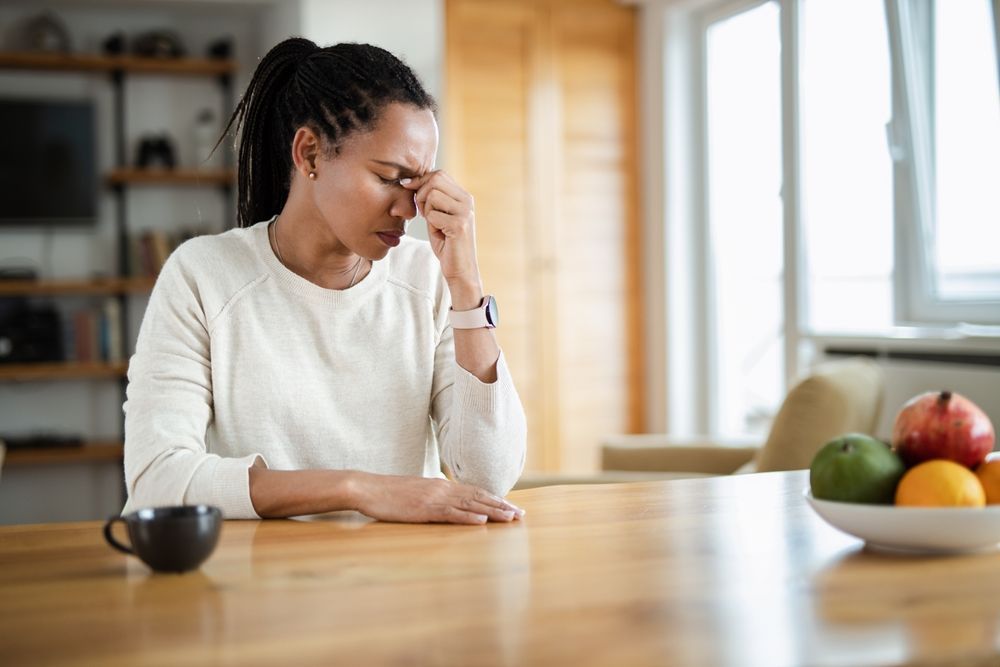Woman at a kitchen table rubbing her temple, with a bowl of fruit on the counter and a coffee cup nearby
