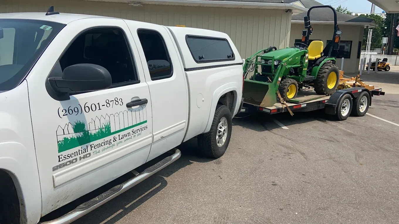White pickup truck towing a flatbed trailer carrying a green tractor in a parking lot