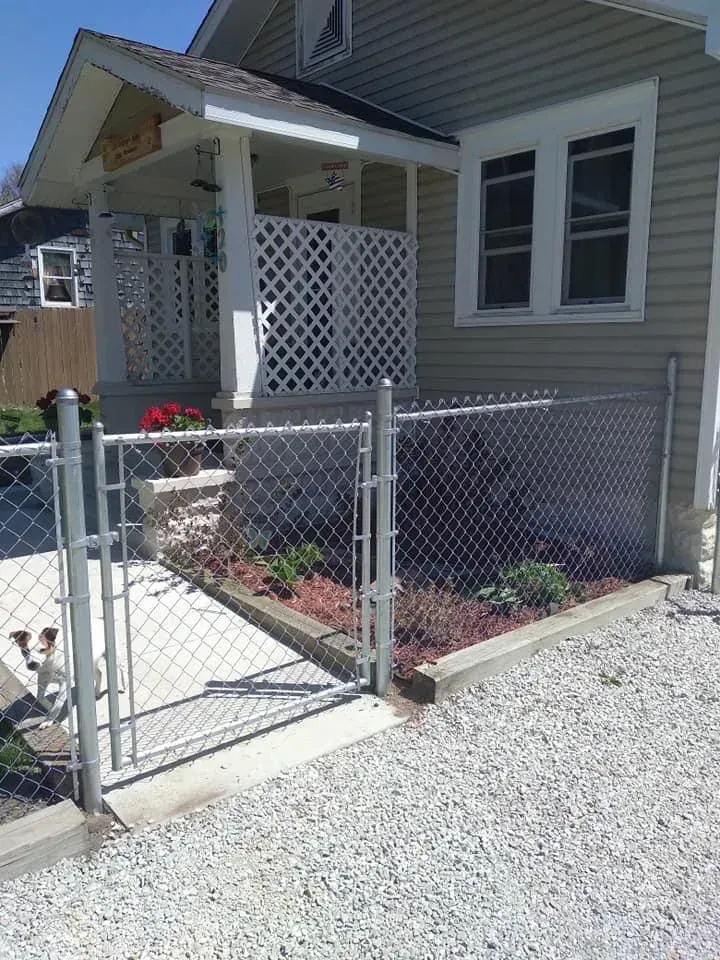 Small front porch and walkway with a chain-link gate, gravel yard, and house windows.