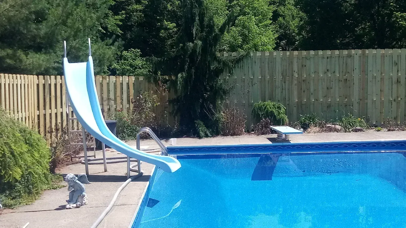 Backyard pool with blue-and-white slide beside a wooden fence and greenery