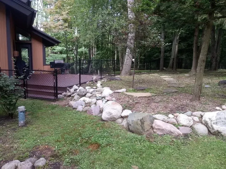 Rocky garden border beside a brown house, with a small deck and wooded backyard.