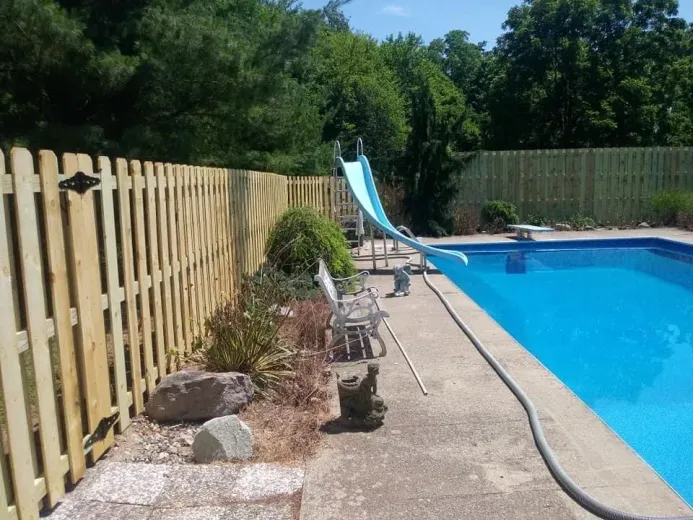 Backyard swimming pool beside a wooden fence, with lounge chairs and a diving board under sunny trees