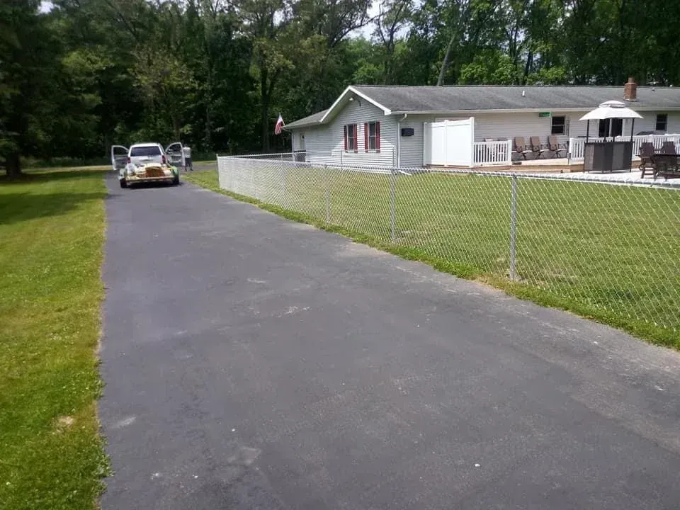 Driveway leading to a white house with a chain-link fence and a parked car on the left.