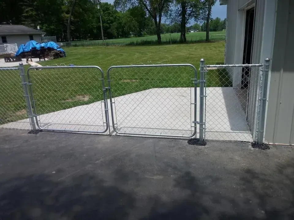 Chain-link gate beside a gray building, opening onto a concrete patio and grassy park.